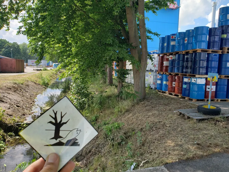 Eine Hand hält das Gefahrensymbol für gewässergefährdende Stoffe vor ein Fasslager, im Vordergrund ein Wassergraben