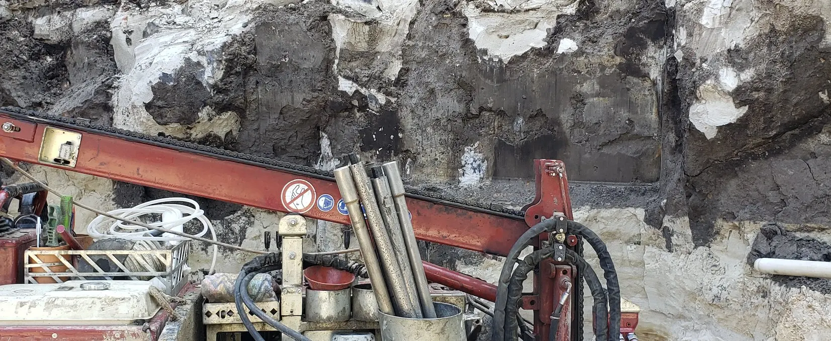 Bohrgerät mit Gestänge vor der aufgeschlossenen Wand einer Baugrube, dunkle und helle Bodenschichten sichtbar